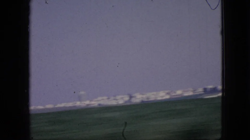 1967 Iowa 8mm frame with a tilted moving-vehicle view across green fields toward a distant city edge or low skyline on the horizon.