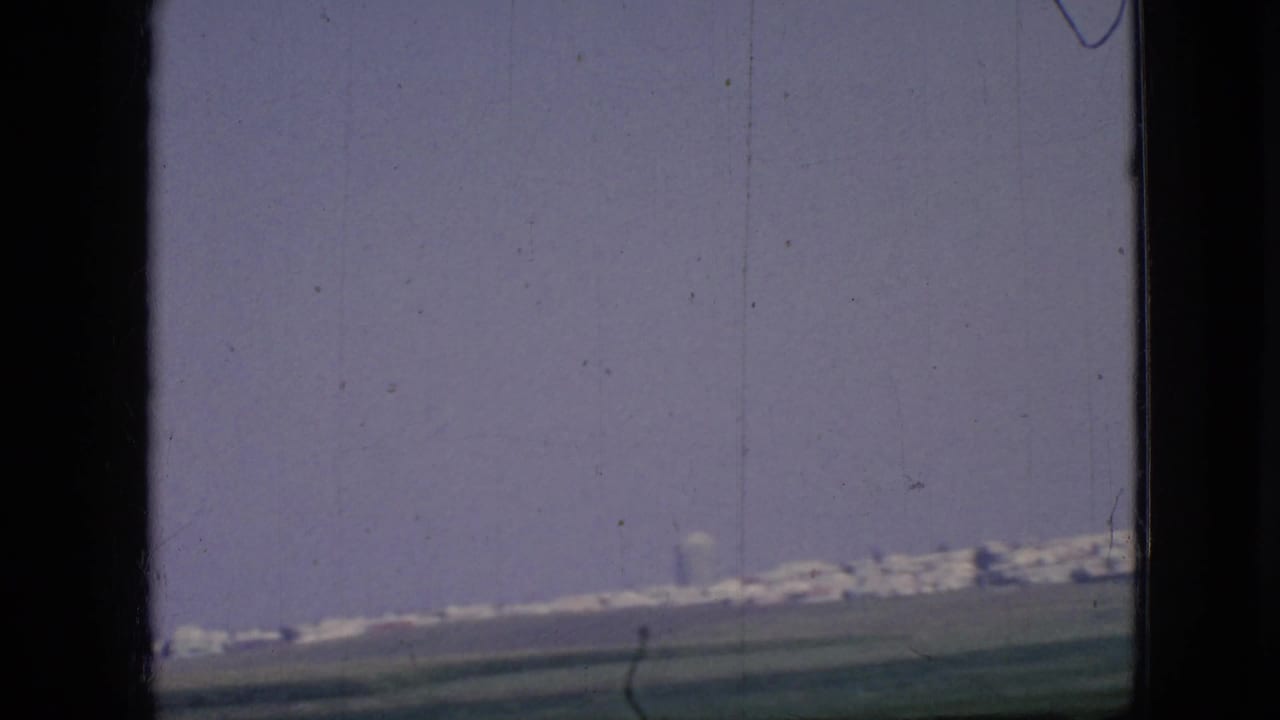 1967 Iowa home-movie frame showing green fields, roadside power lines, and a distant city edge on the horizon through soft 8mm color.
