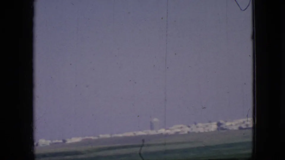 1967 Iowa home-movie frame showing green fields, roadside power lines, and a distant city edge on the horizon through soft 8mm color.