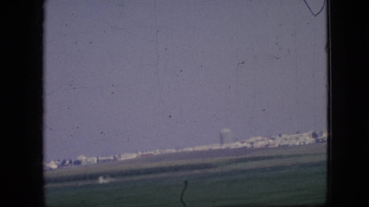1967 Iowa 8mm home-movie frame showing open rural fields and a distant line of low buildings viewed through a dark vehicle window frame.