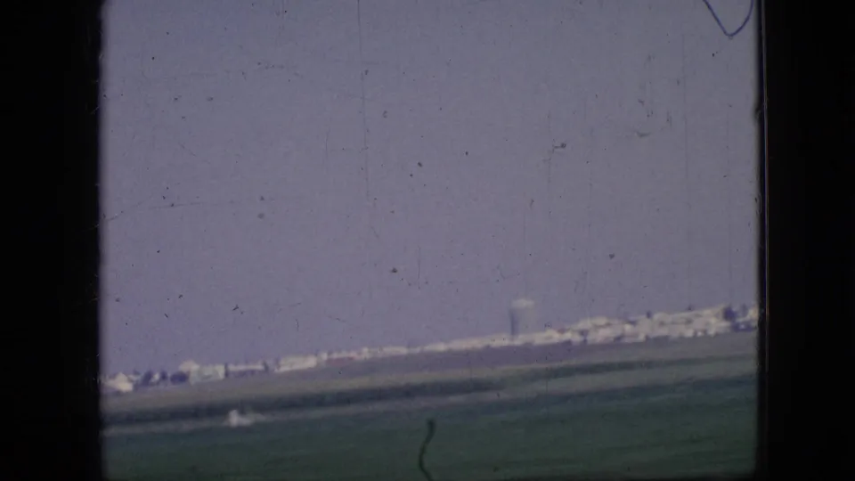 1967 Iowa 8mm home-movie frame showing open rural fields and a distant line of low buildings viewed through a dark vehicle window frame.