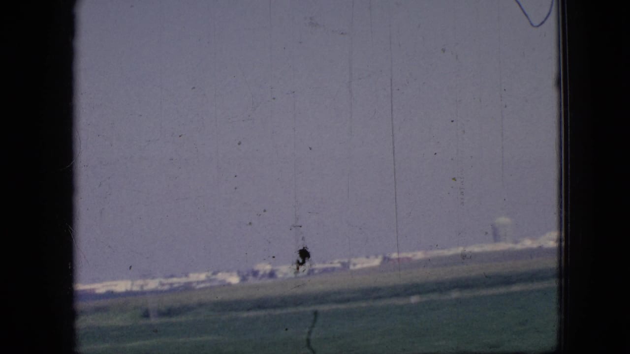 1967 Iowa road-travel frame with distant low buildings beyond flat farmland, seen through dark vertical window or door framing on scanned 8mm film.