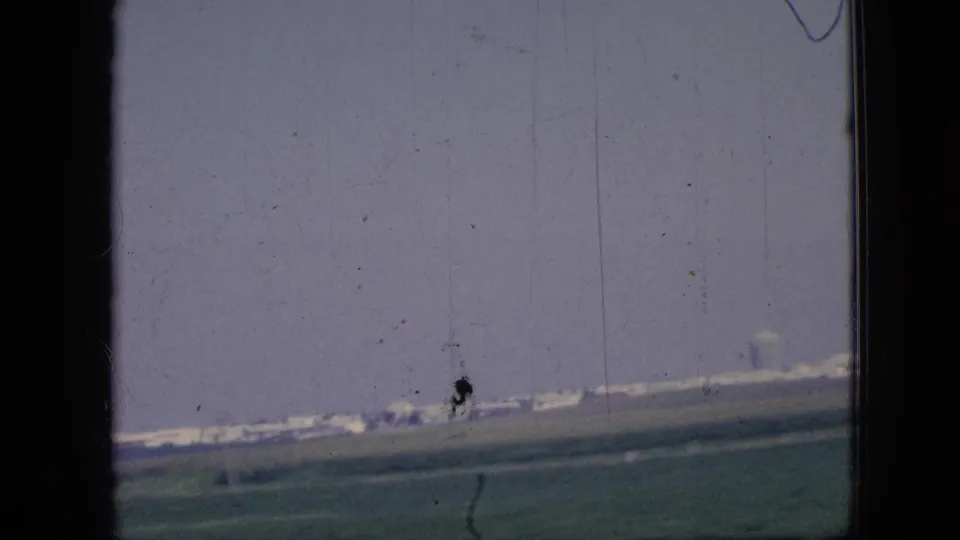 1967 Iowa road-travel frame with distant low buildings beyond flat farmland, seen through dark vertical window or door framing on scanned 8mm film.