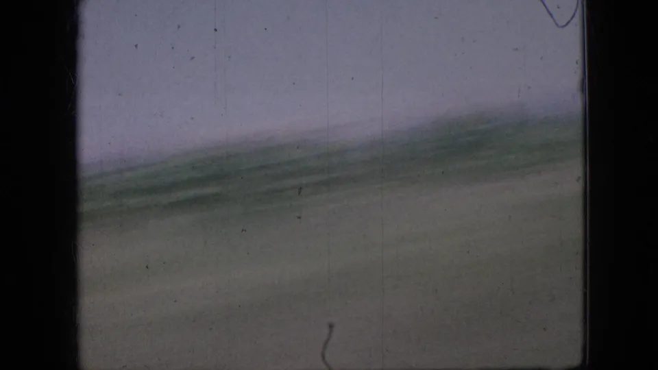 1967 Iowa home-movie frame with motion-blurred fields and a sloping horizon, emphasizing the passenger view from a moving road vehicle.