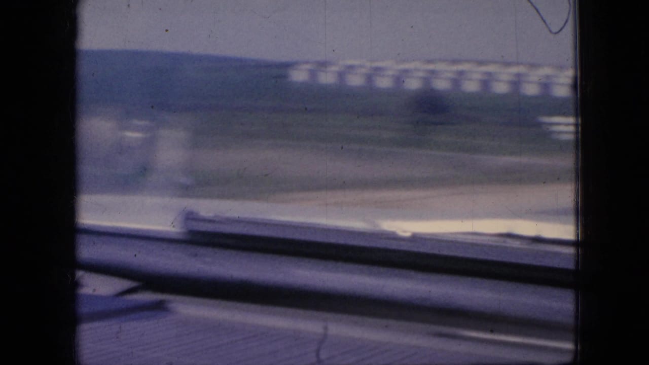 1967 Iowa 8mm frame from a moving car showing a blurred rural highway edge, dashboard or windshield detail, grassy roadside, and distant white silos.