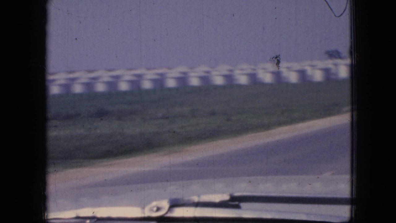 Roadside curve with a row of white silos beyond the grass and windshield hardware at the bottom.