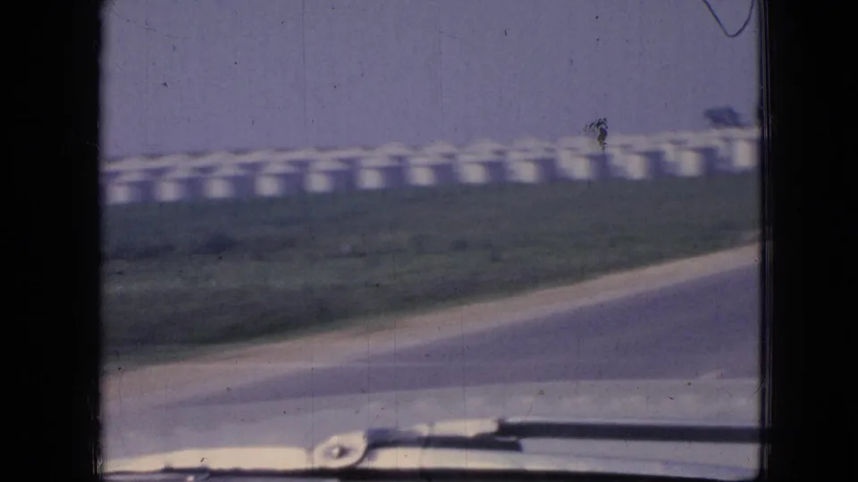 1967 Iowa home-movie frame showing a curving roadside, low green verge, distant white agricultural silos, and the lower edge of a car windshield.