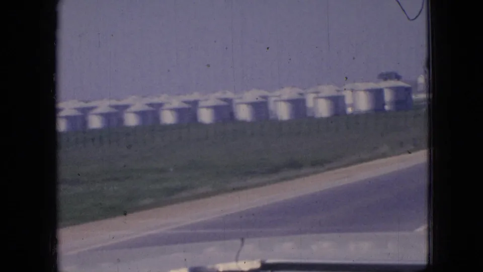 1967 Iowa 8mm frame with strong motion blur as the car passes a grassy roadside and a line of white silos under an open sky.