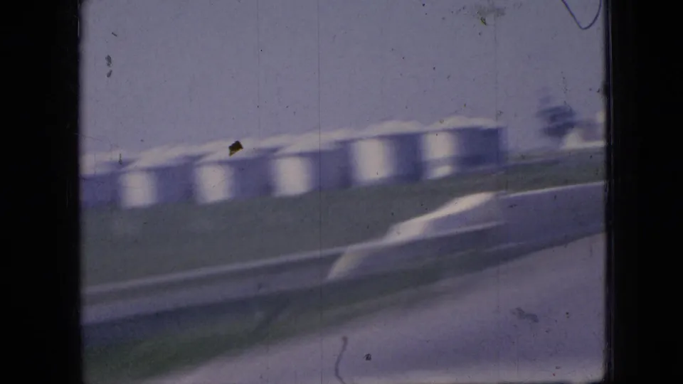 1967 Iowa home-movie frame showing a moving-car view of distant white silos, a paved road, grassy verge, and visible 8mm film wear.
