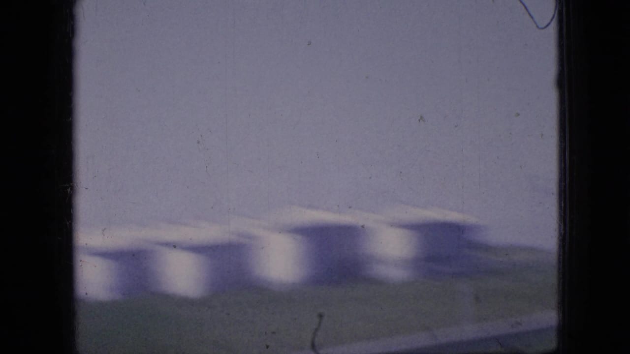 1967 Iowa 8mm frame showing soft, motion-blurred white grain silos across open land, partly obscured by the dark vertical edge of a car window.