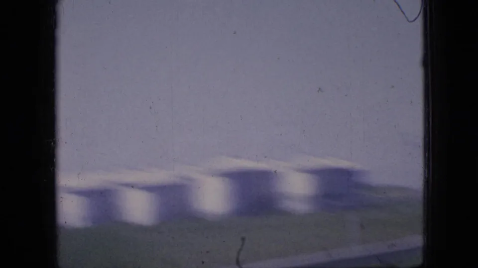 1967 Iowa 8mm frame showing soft, motion-blurred white grain silos across open land, partly obscured by the dark vertical edge of a car window.