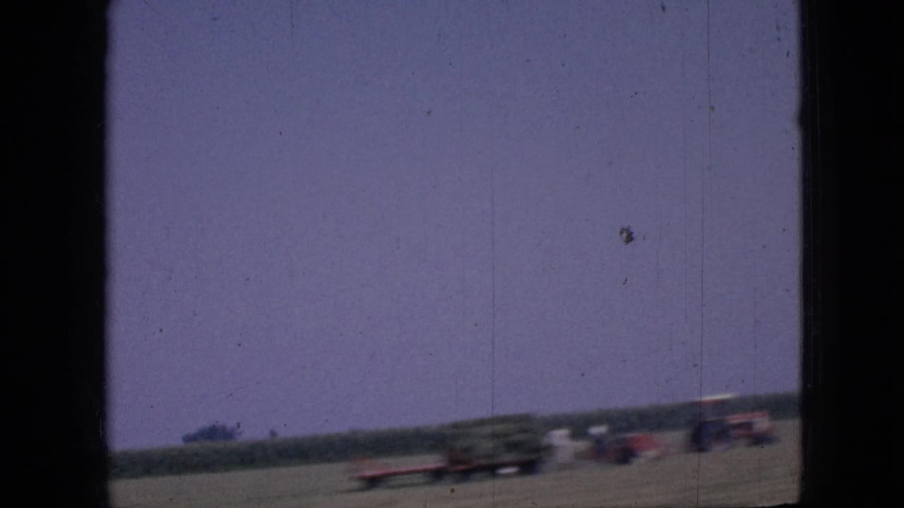 A moving-vehicle view in 1967 Iowa shows blurred cars or tractors along a flat roadside beneath a purple-tinged sky and distant tree line.