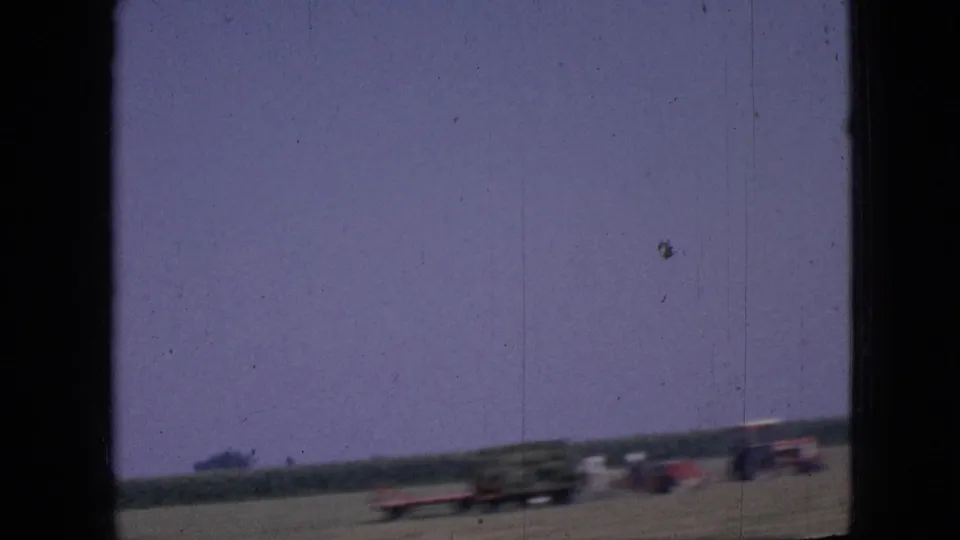A moving-vehicle view in 1967 Iowa shows blurred cars or tractors along a flat roadside beneath a purple-tinged sky and distant tree line.