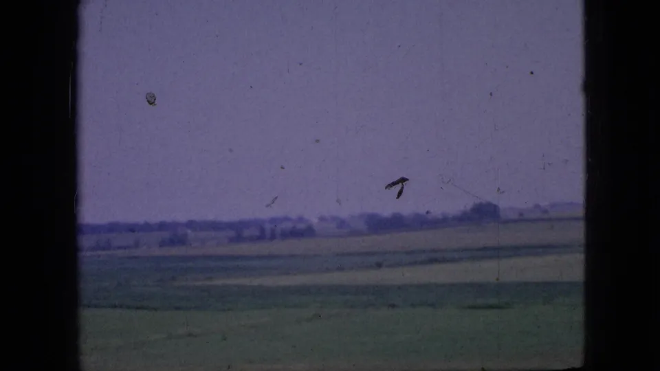 A 1967 Iowa countryside frame shows broad green and tan fields, a low distant tree line, and a pale purple sky with visible film scratches.