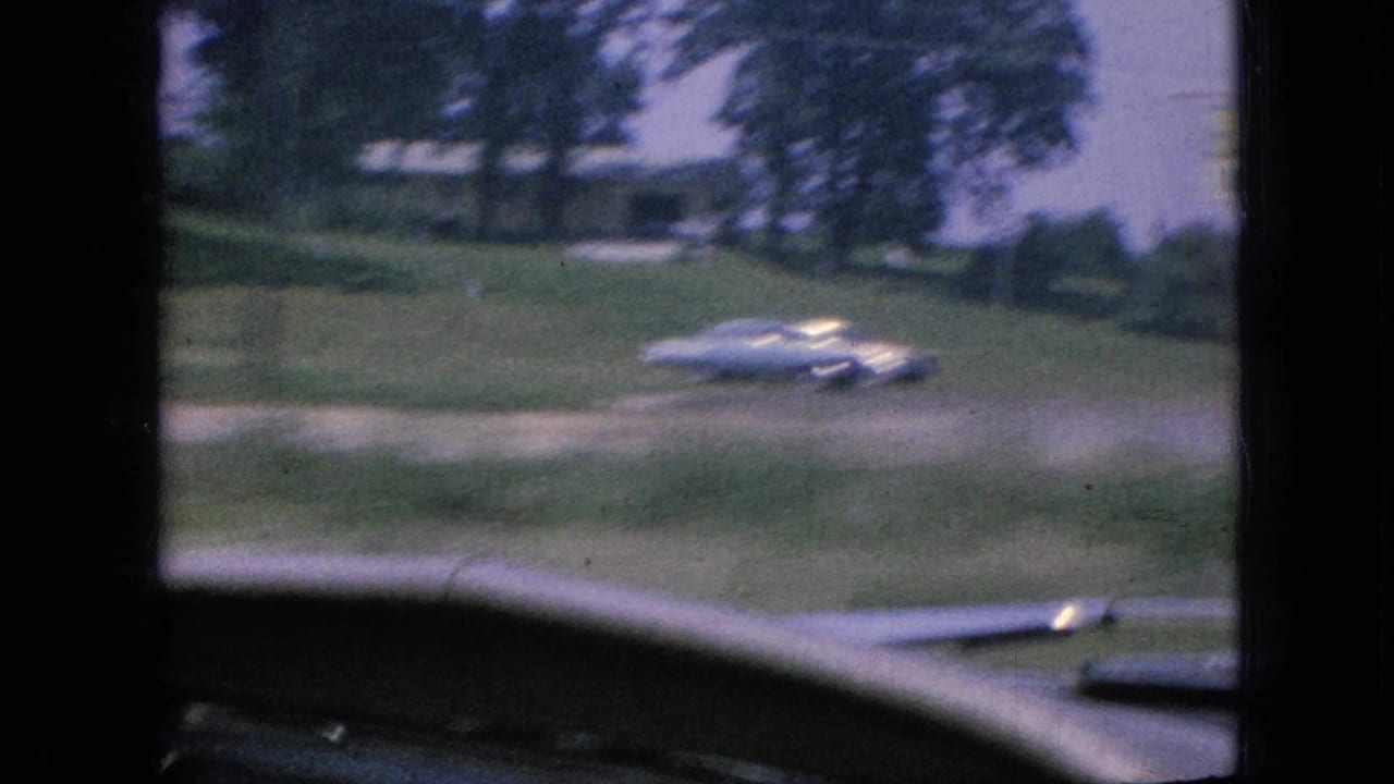 1967 Iowa home-movie frame looking through a moving car windshield toward a grassy roadside, trees, and a white mid-century car.