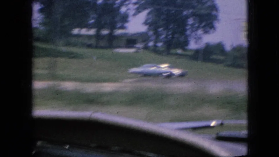 1967 Iowa home-movie frame looking through a moving car windshield toward a grassy roadside, trees, and a white mid-century car.