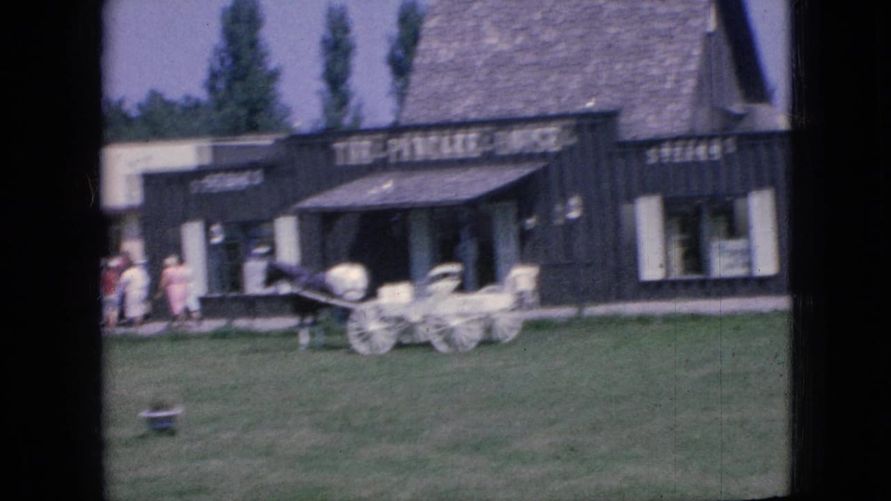 1967 Iowa roadside stop seen from a moving car, with a dark building, several people near the entrance, and a horse-drawn carriage display on the lawn.