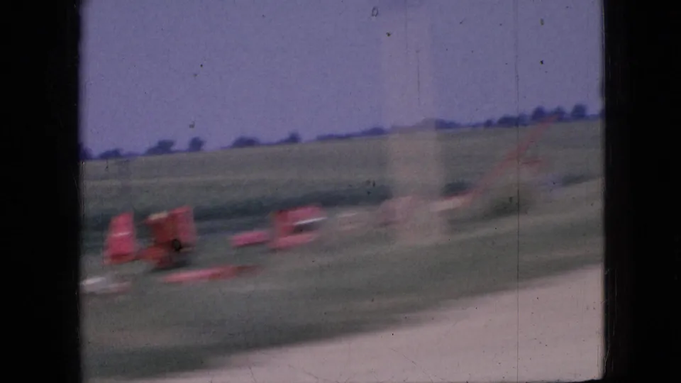 1967 Iowa home-movie frame with motion-blurred red roadside structures, open fields, horizon trees, and the car window edge visible during travel.