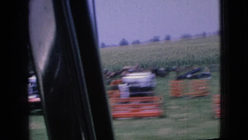 1967 Iowa moving-car view framed by the vehicle interior, showing a rural field, red fencing or equipment, and a light-colored car or object near the roadside.