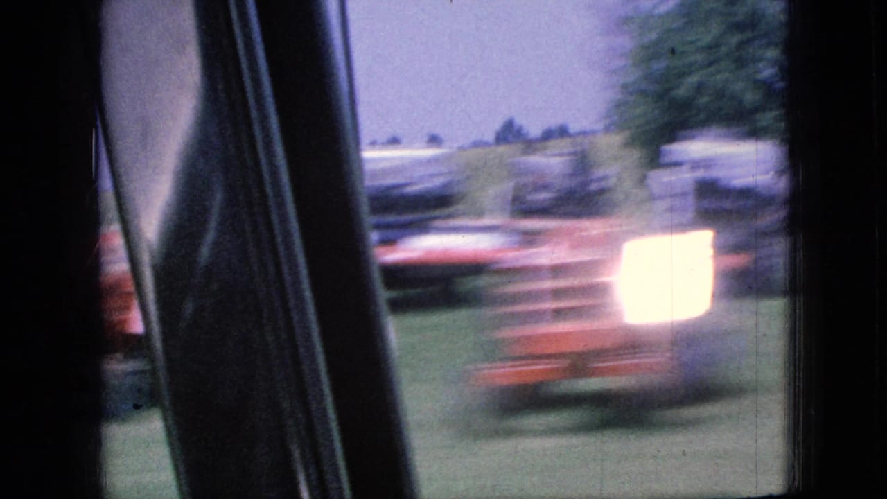 1967 Iowa 8mm frame looking through a moving car window at a blurred roadside area with dark interior trim, grass, trees, and distant vehicles or structures indistinct in motion.