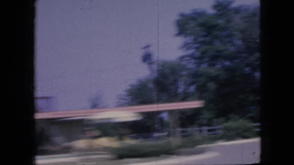1967 Iowa road-trip frame showing a blurred roadside building, trees, and dark vehicle-window framing as the camera moves past the scene.