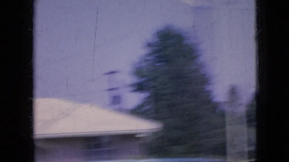 1967 Iowa 8mm frame with a pale roofline, roadside trees, and open sky viewed from a moving vehicle, with heavy blur and faded color.