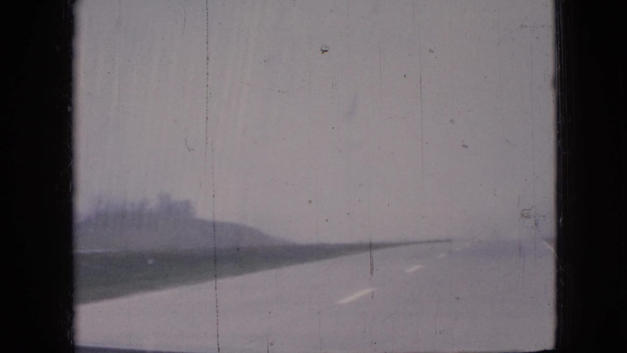 1967 Iowa roadside landscape seen through a dusty vehicle window, with a flat road edge and hazy overcast sky.
