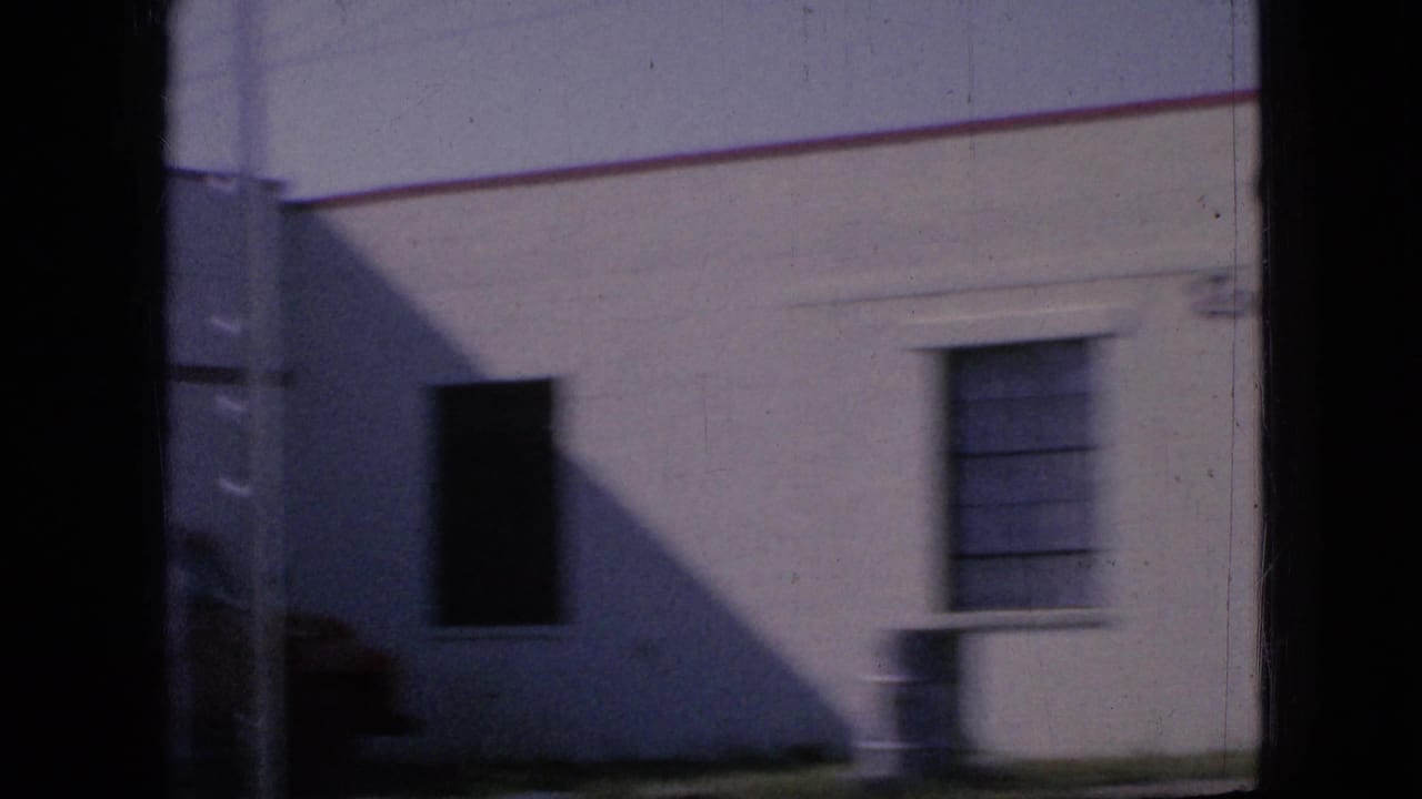 1967 Iowa home-movie frame showing a white roadside building with rectangular windows, seen obliquely through a moving vehicle with dark film or car-frame edges.