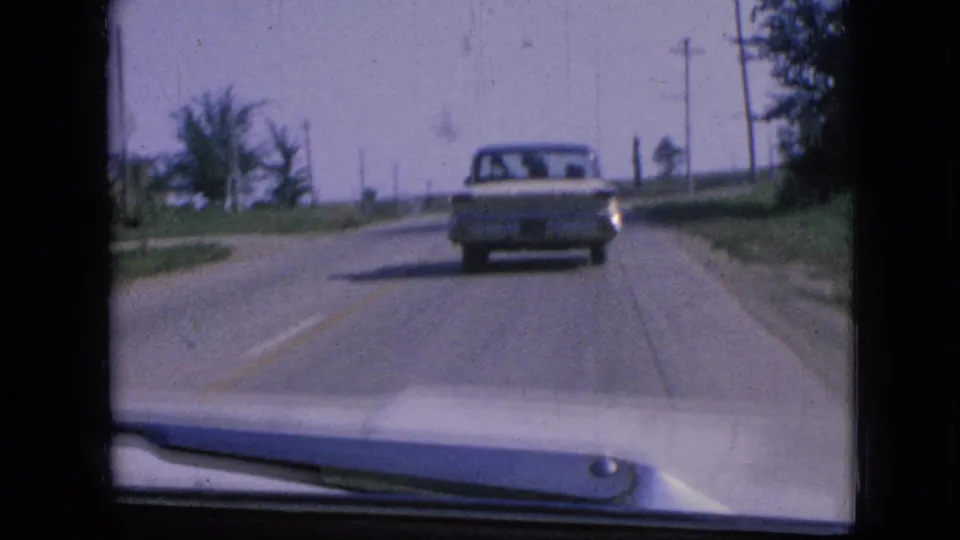 1967 Iowa road view through a windshield, with a mid-century car ahead on a two-lane road bordered by utility poles and roadside greenery.