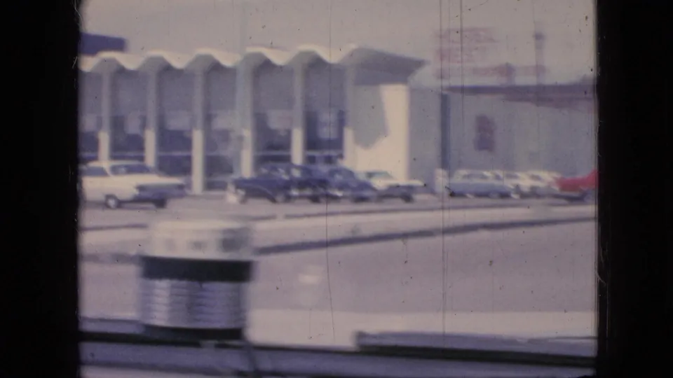 1967 Iowa moving-car frame showing a white building with repeated arched bays, parked cars along the frontage, and windshield elements in the foreground.