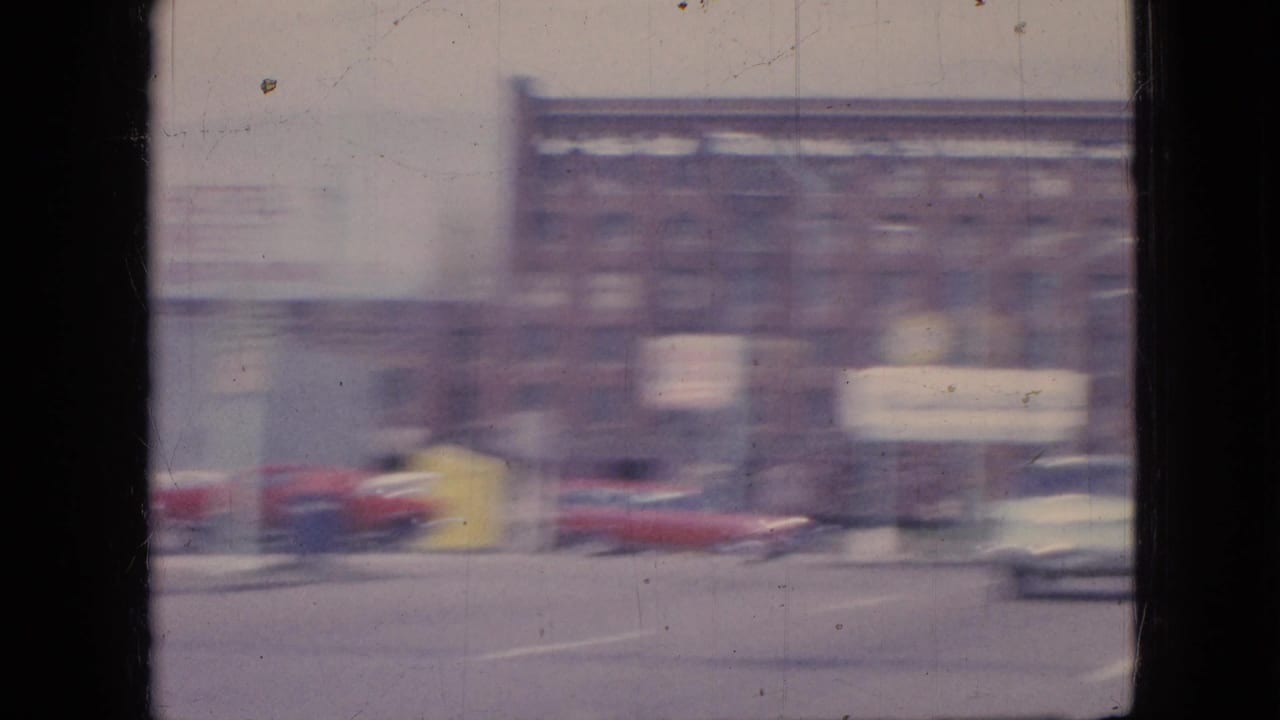 1967 Iowa home-movie frame showing a blurred mid-century street with older cars, road markings, brick commercial buildings, and a pale sky.