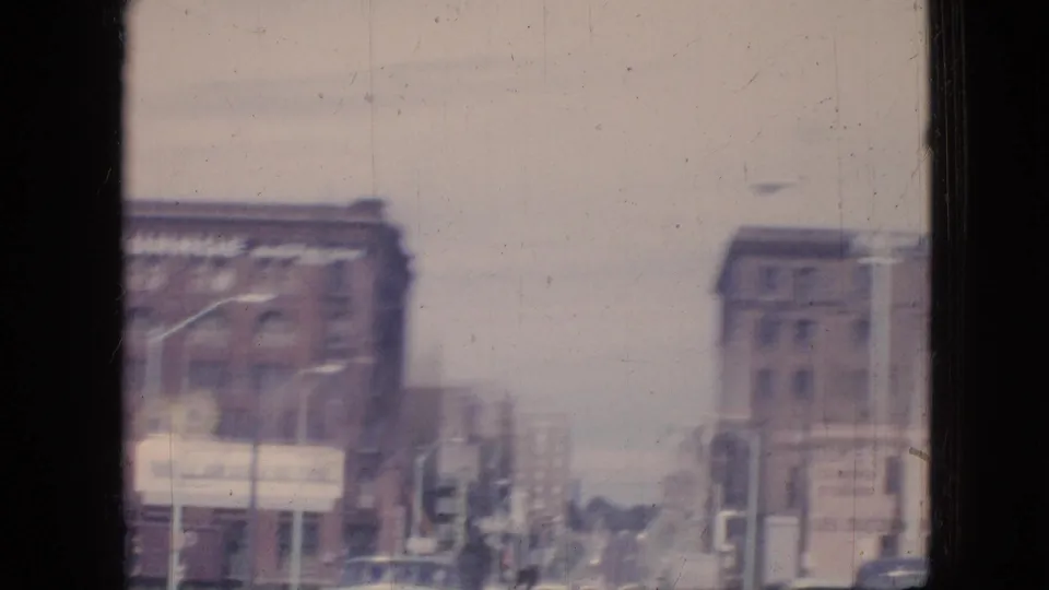 1967 Iowa street view with brick buildings, streetlights, older vehicles, and a prominent storage sign visible through soft 8mm focus.