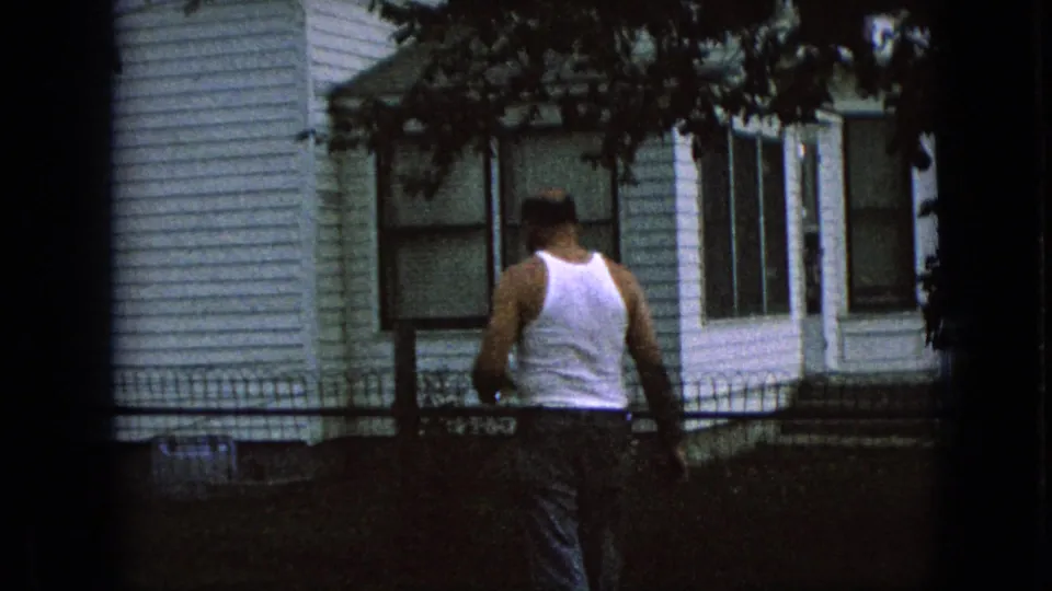 1967 Iowa residential frame showing a man in a white tank top near a white-sided house, dark fence, shaded lawn, and overhanging tree branches.
