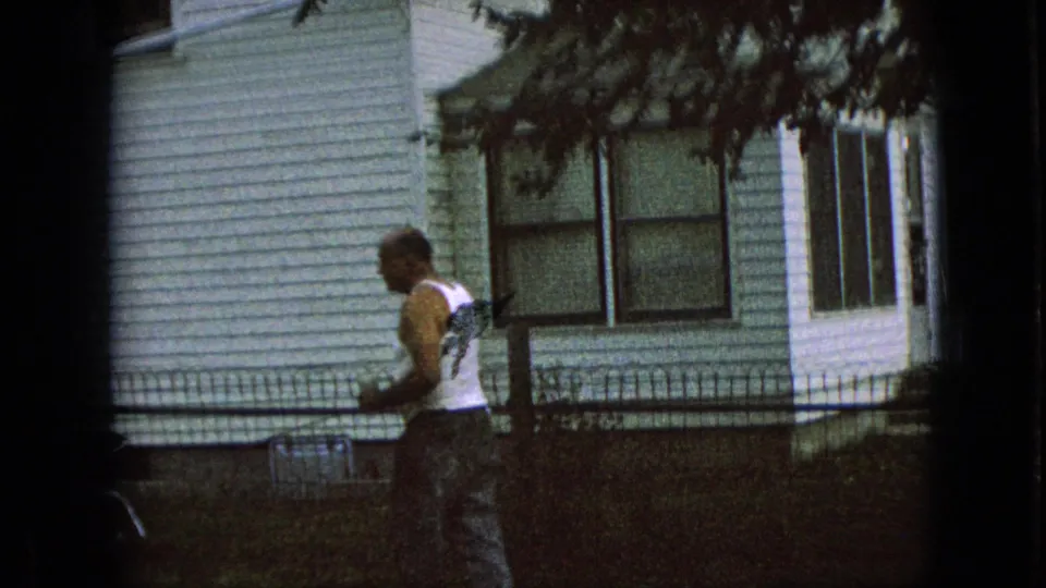 1967 Iowa home-movie frame of a man walking in a shaded residential yard beside a white-sided house with windows and a low fence.
