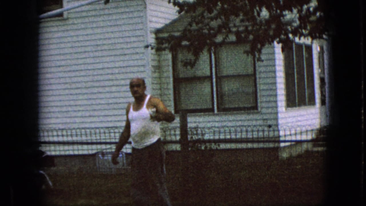 In supplied 1967 Iowa home-movie footage, an adult man in a white tank top walks across a residential yard with a white-sided house and low fence behind him.
