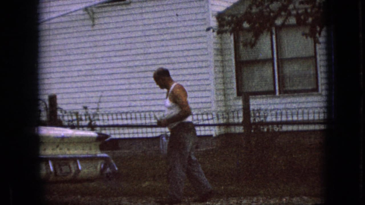 Man holding a drink walks near a pale parked car in front of a house.