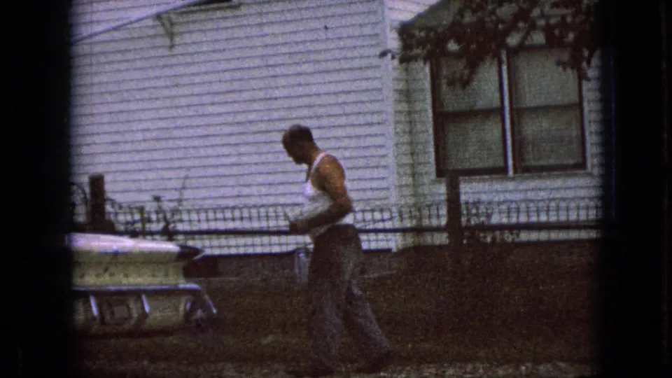The man appears to hold a drink while moving beside a pale parked car, with the white house and fence visible in the background.