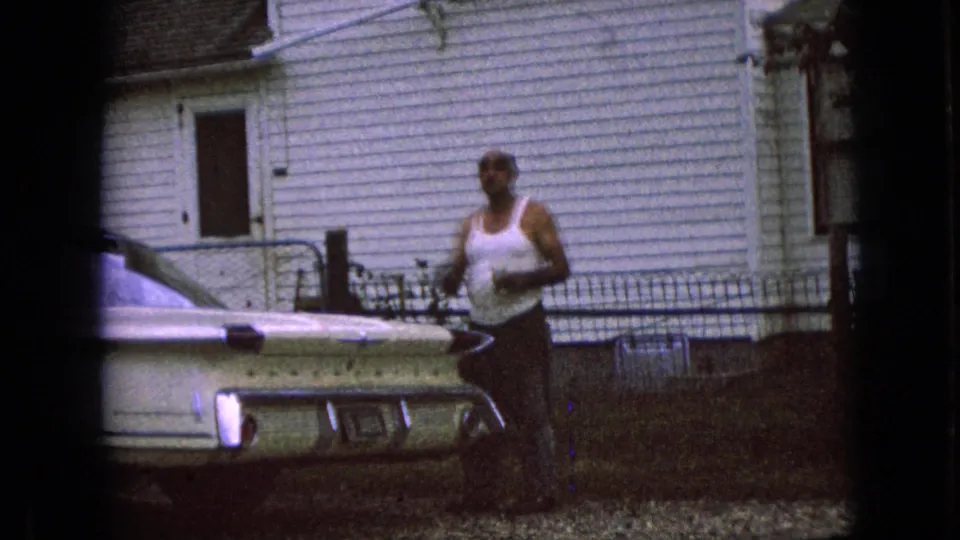 The man pauses or walks near the rear of a pale classic car in a residential yard, with house siding, windows, and fencing behind him.