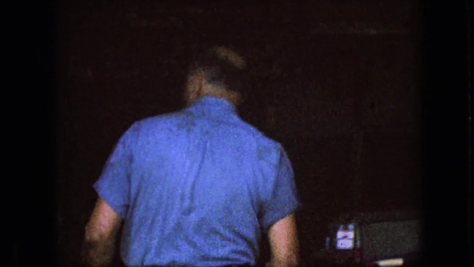A second adult man in a blue short-sleeve shirt is seen from behind in a darker home-movie frame, with limited surrounding detail visible.