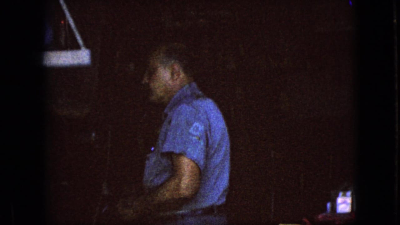 In this 1967 Iowa 8mm home-movie frame, an older man in a blue short-sleeved shirt stands in profile or partly turned away inside a dim garage workspace.