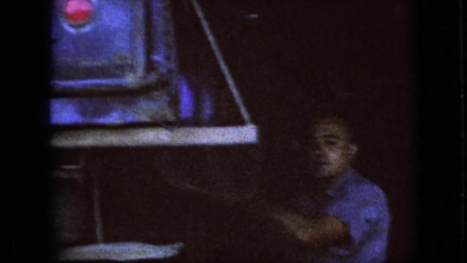 The man appears beside a shelf or bench in the dark garage, with part of a large object or machine visible above him in the 1967 Iowa home-movie footage.