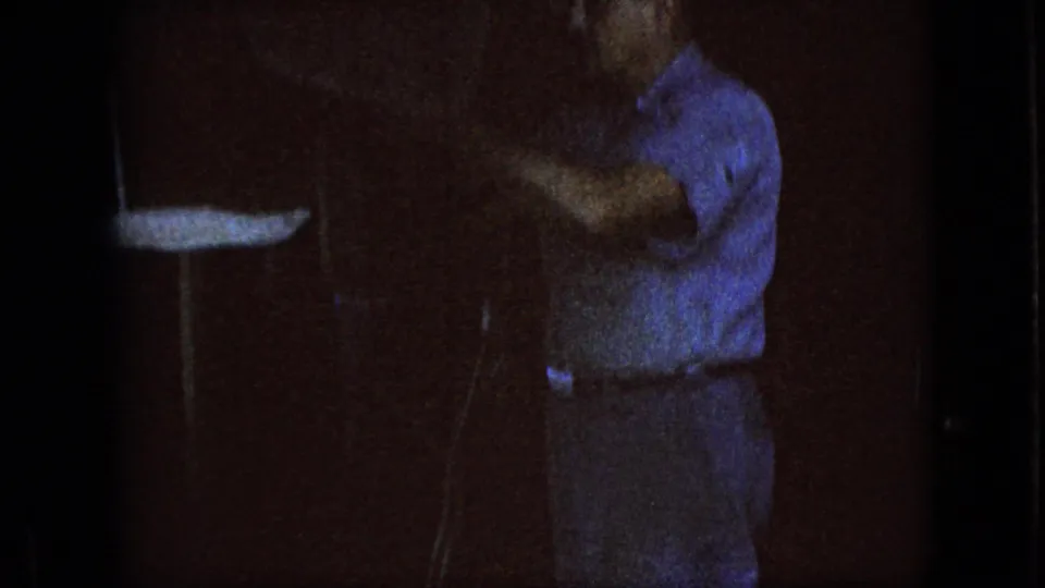 The older man is seen from the side in a low-light garage, raising an arm near a work surface or hanging fixture in the supplied 1967 Iowa home-movie clip.