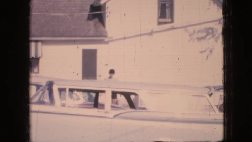 1967 Iowa 8mm frame showing a white station wagon parked along a pale residential building while a woman walks near the car in bright daylight.