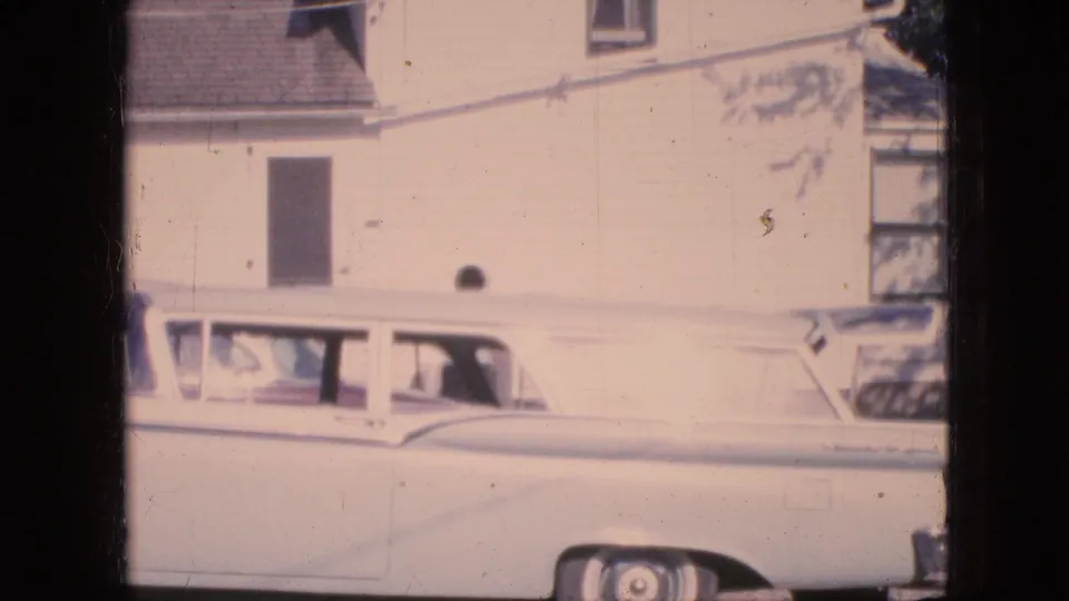 1967 Iowa home-movie frame showing the parked white station wagon and pale residential facade, with a person visible in or near the vehicle area.