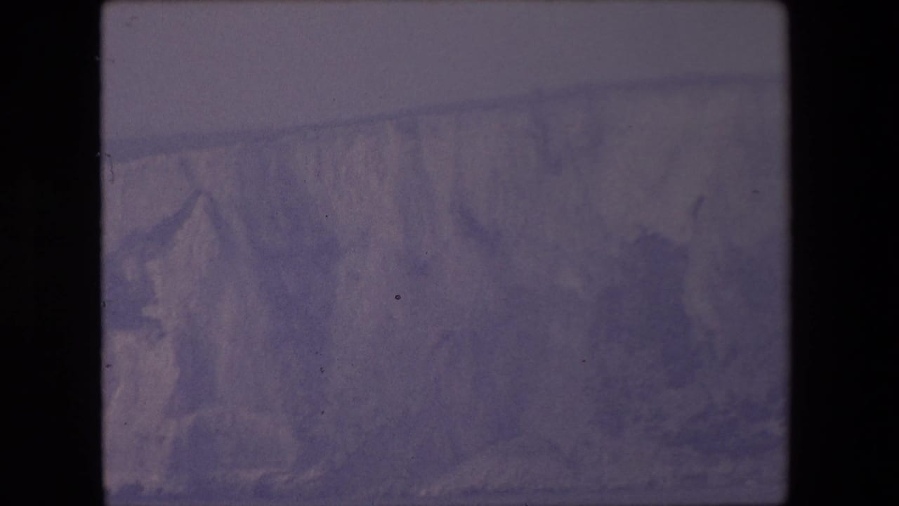 Purple-tinted 8mm frame of a distant snowy or icy mountain face behind dark film borders.