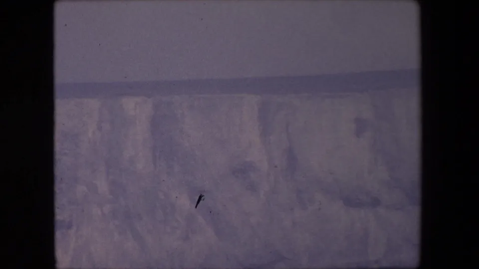 1967 home-movie frame showing a pale rocky or icy slope with canyon-like depth, visible dust specks, soft exposure, and heavy film grain.