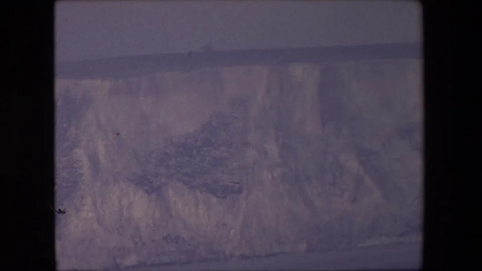 1967 8mm frame showing a steep mountain face with snow- or ice-like textures, a muted horizon, purple color shift, and rounded film gate edges.