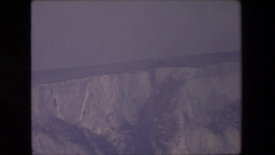 1967 8mm home-movie frame with the camera lowered toward a pale canyon wall, distant rim line, and washed, grainy sky.