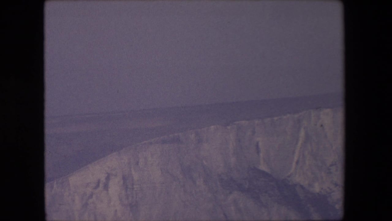 1967 8mm home-movie frame from supplied London, England metadata showing a distant pale cliff face, muted sea, and overcast sky.