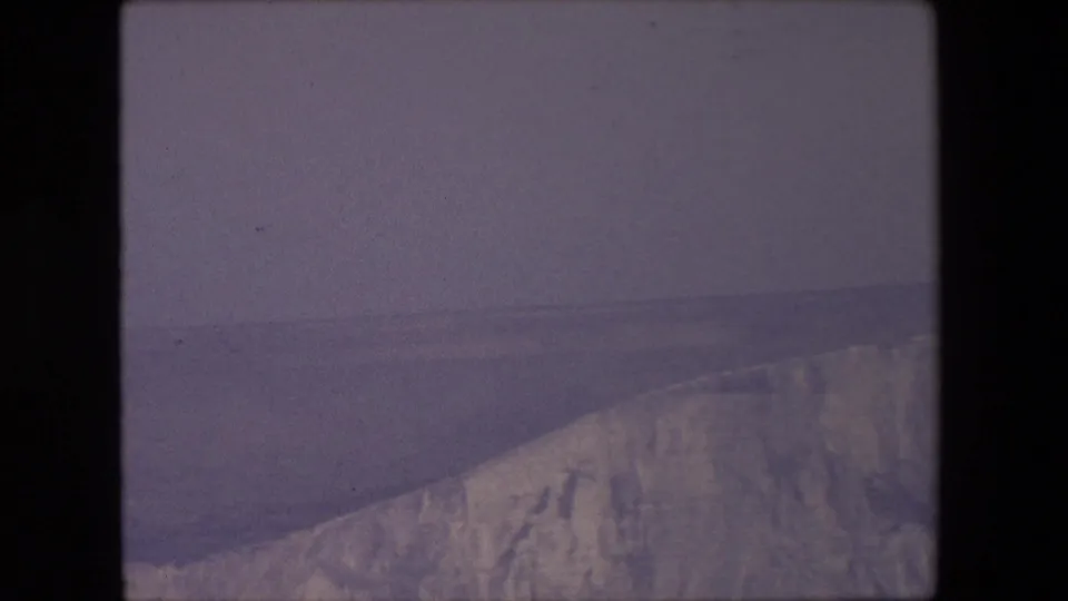 1967 8mm frame showing a rugged pale cliff ridge across calm sea, with muted color and soft home-movie focus.